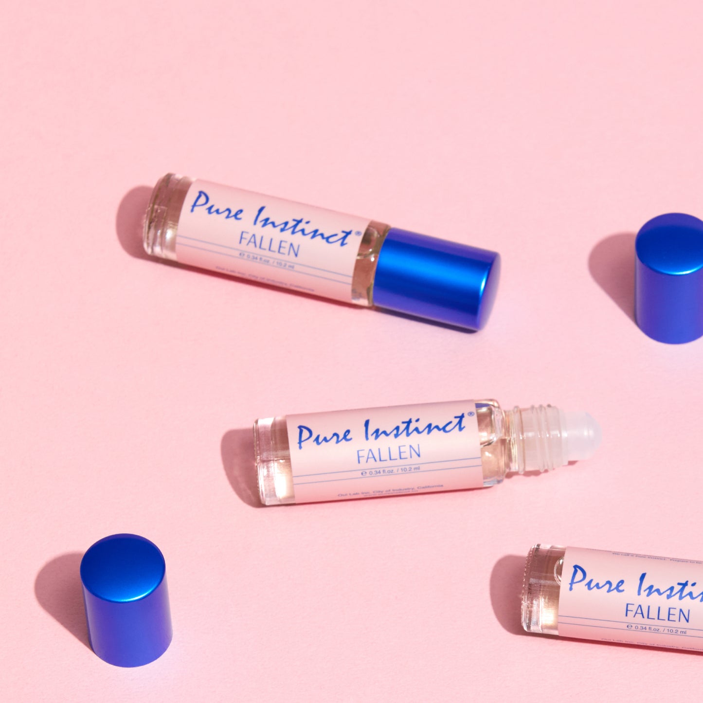 Pure Instinct FALLEN roll-on perfume oil bottles with blue caps arranged on a soft pink background