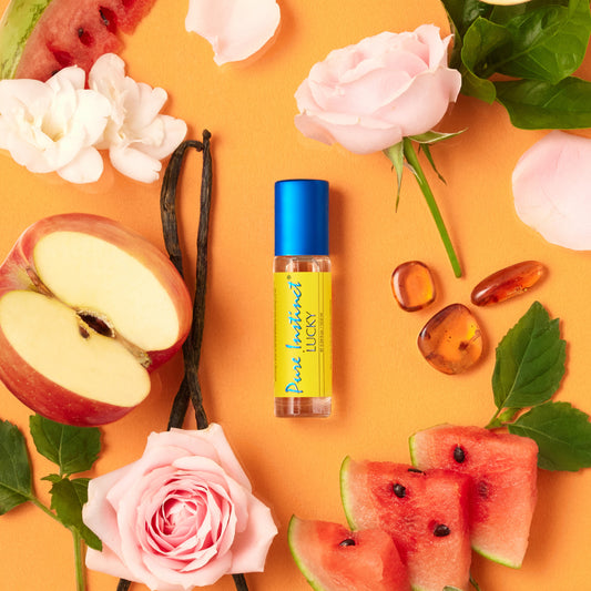 Lucky Roll-On Pheromone Perfume Oil surrounded by roses, apple slices, watermelon, vanilla beans, and amber stones on a warm peach background.