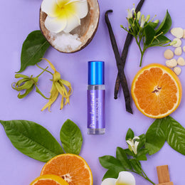 Surrender Roll-On Pheromone Perfume Oil surrounded by orange slices, vanilla pods, ylang-ylang, coconut flakes, white flowers and greenery, showcasing its warm and tropical scent profile