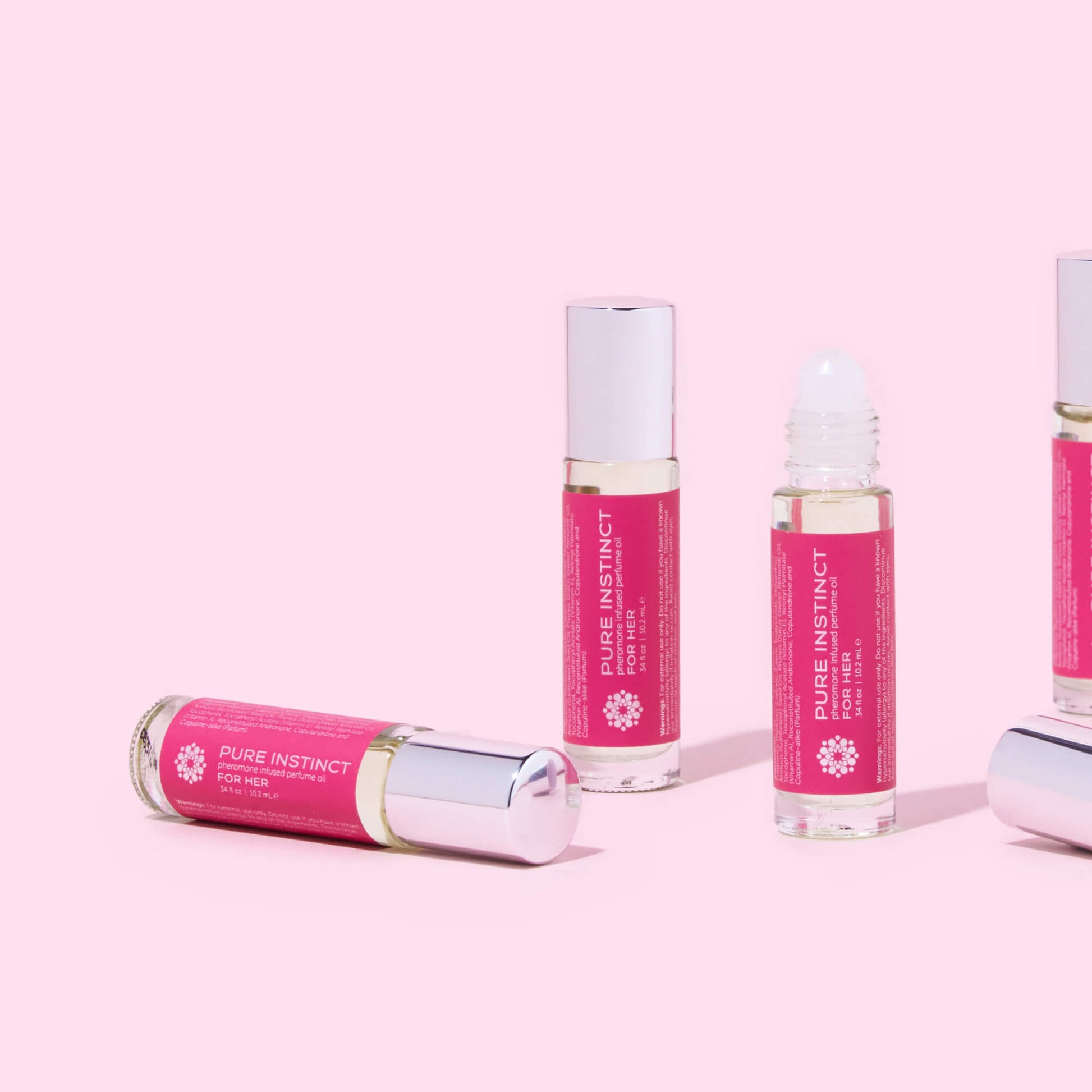 Multiple bottles of FOR HER Roll-On Pheromone Perfume Oil arranged on a soft pink background, featuring the sleek silver cap and signature magenta label design.