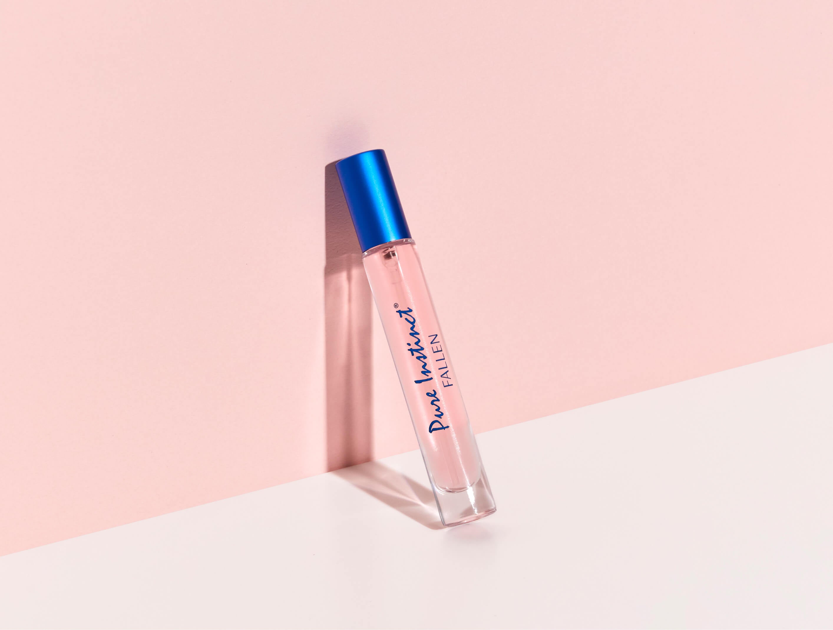 Pure Instinct Fallen pheromone perfume spray standing against a soft pink wall with a bold blue cap and light pink fragrance inside