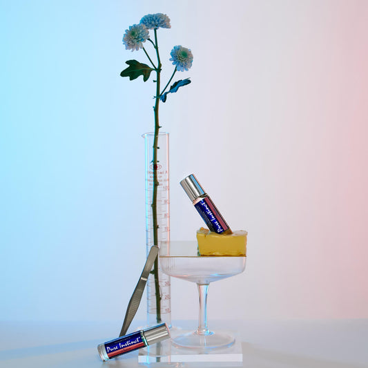 Pure Instinct Crave Roll-On Pheromone Perfume Oil displayed creatively with a sliced butter, glassware, and a single stem flower under soft blue and pink lighting — evoking a playful, tropical-lab mood.