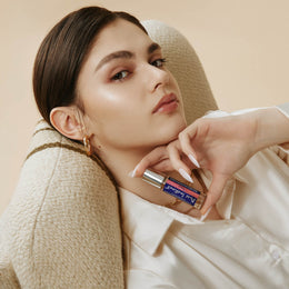 A relaxed woman lounges in a cozy chair while holding Pure Instinct Crave Roll-On Pheromone Perfume Oil close to her face, exuding confidence and effortless elegance.