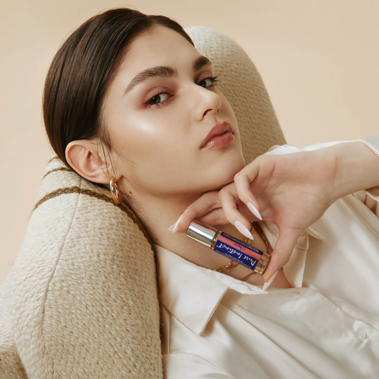 A relaxed woman lounges in a cozy chair while holding Pure Instinct Crave Roll-On Pheromone Perfume Oil close to her face, exuding confidence and effortless elegance.