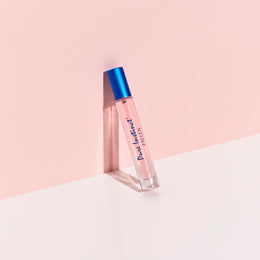 Pure Instinct Fallen pheromone perfume spray standing against a soft pink wall with a bold blue cap and light pink fragrance inside