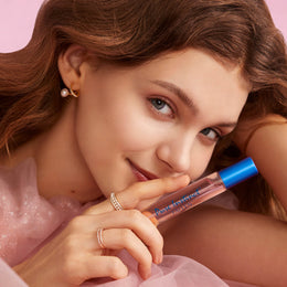 Woman holding Pure Instinct Fallen perfume spray near her lips with a soft smile, wearing pearl and gold jewelry and a sheer pink outfit