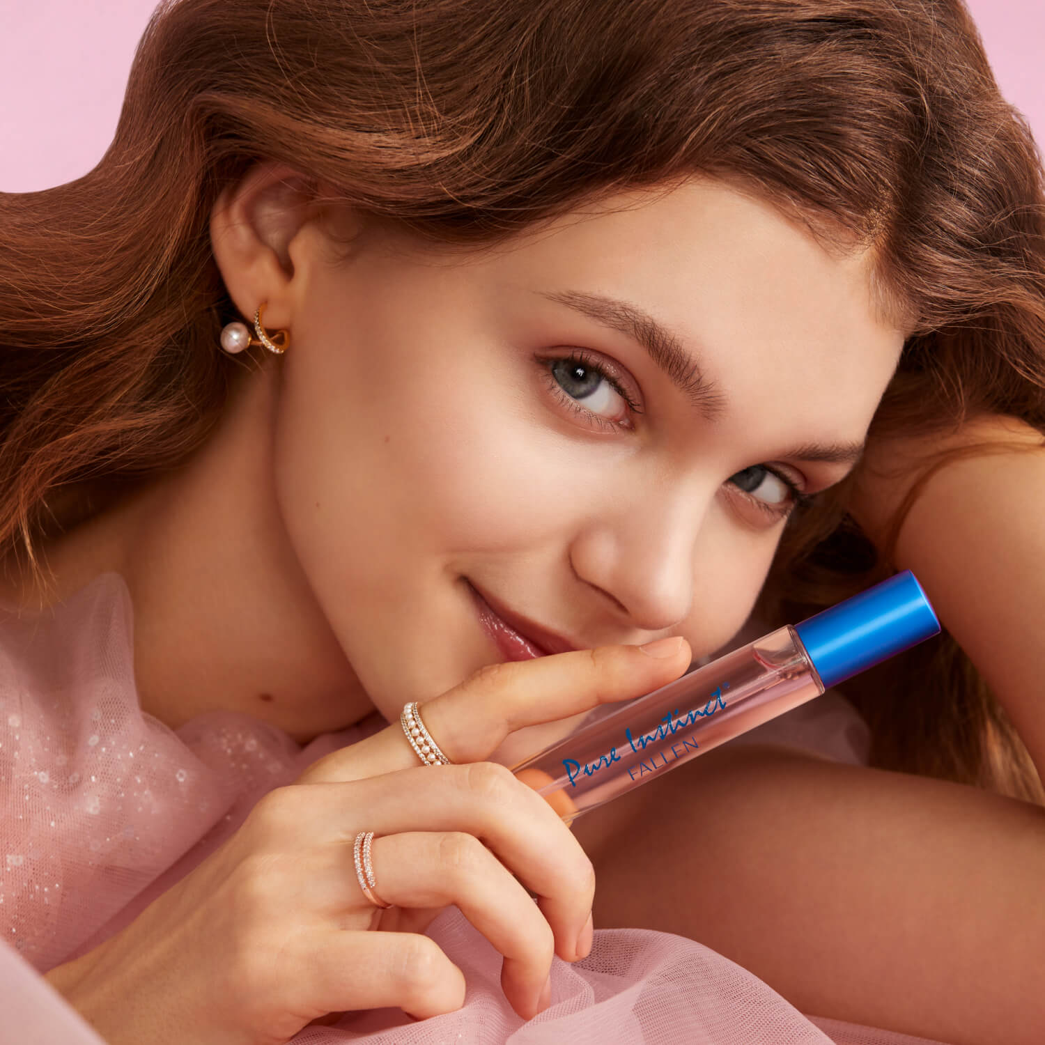 Woman holding Pure Instinct Fallen perfume spray near her lips with a soft smile, wearing pearl and gold jewelry and a sheer pink outfit