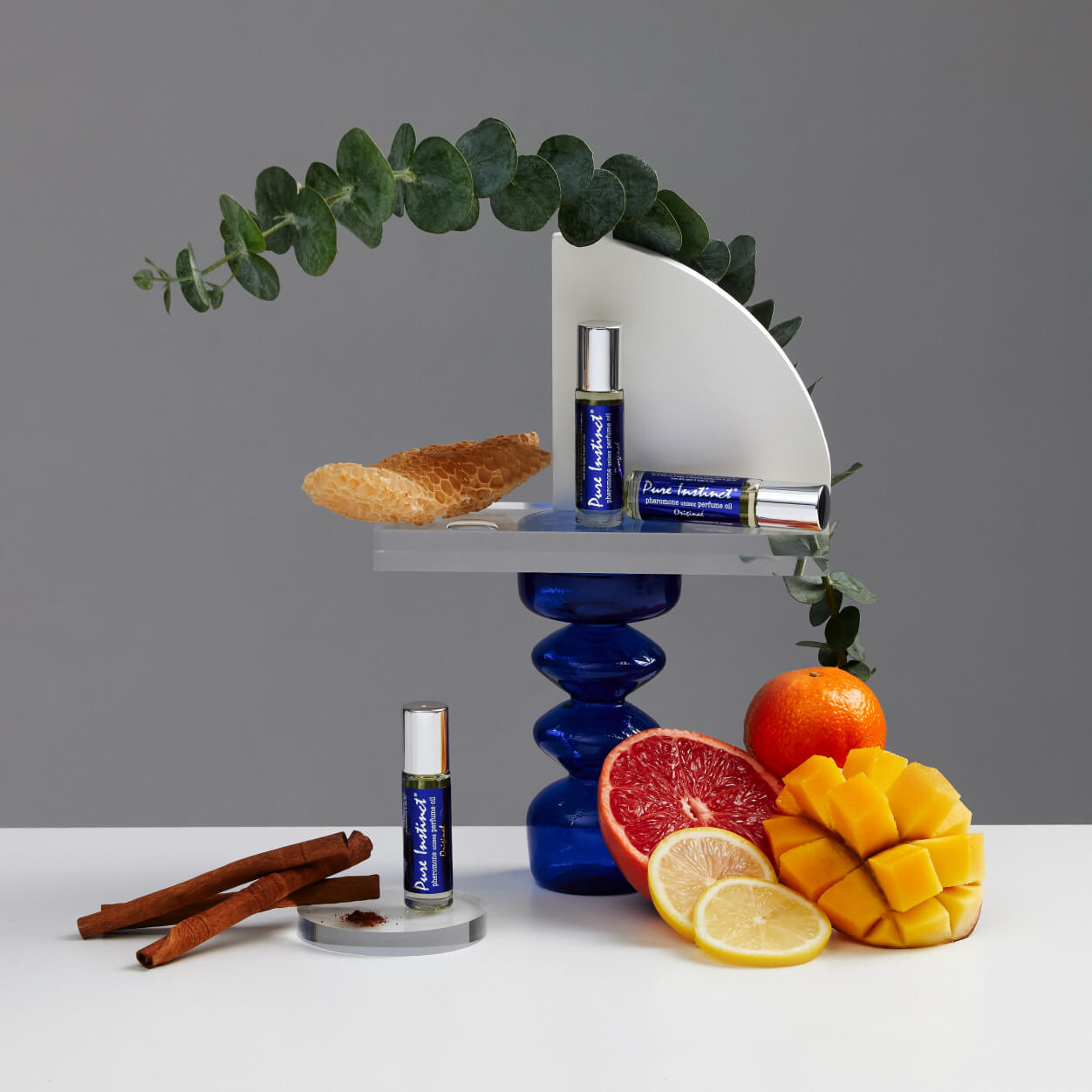 Pure Instinct Original pheromone perfume bottles displayed with citrus fruits, mango, cinnamon, honeycomb, and eucalyptus